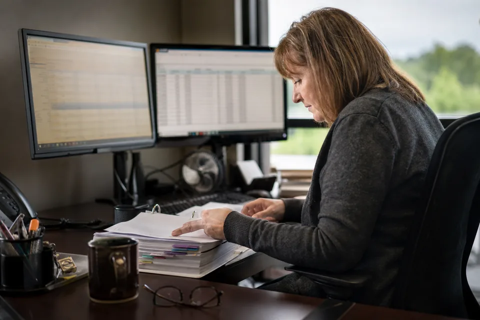 Billing coordinator at a law firm reviewing a rejected electronic invoice on two monitors with a stack of carrier billing guidelines